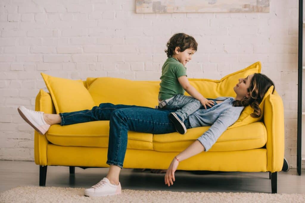 toddler sitting on mom on yellow couch. Mom is exhausted.
