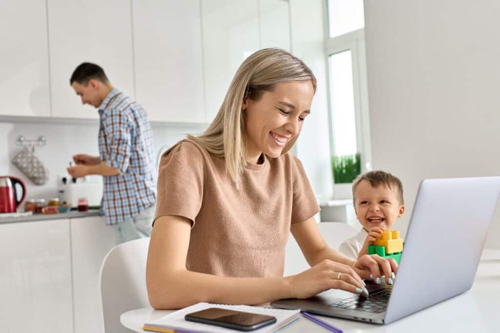 Mom working at kitchen table on laptop with toddler boy next to her. Smiling together.