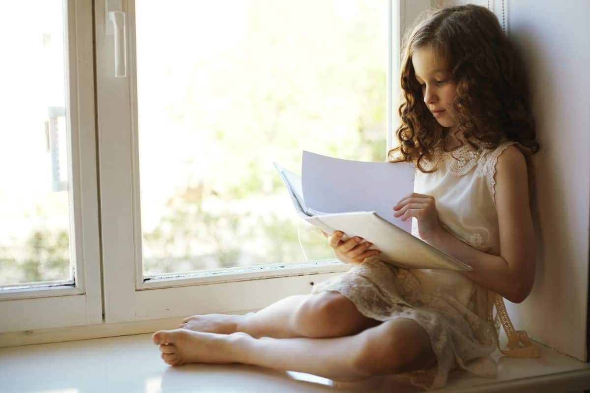 Tween daughter sitting in windowsill reading a book.