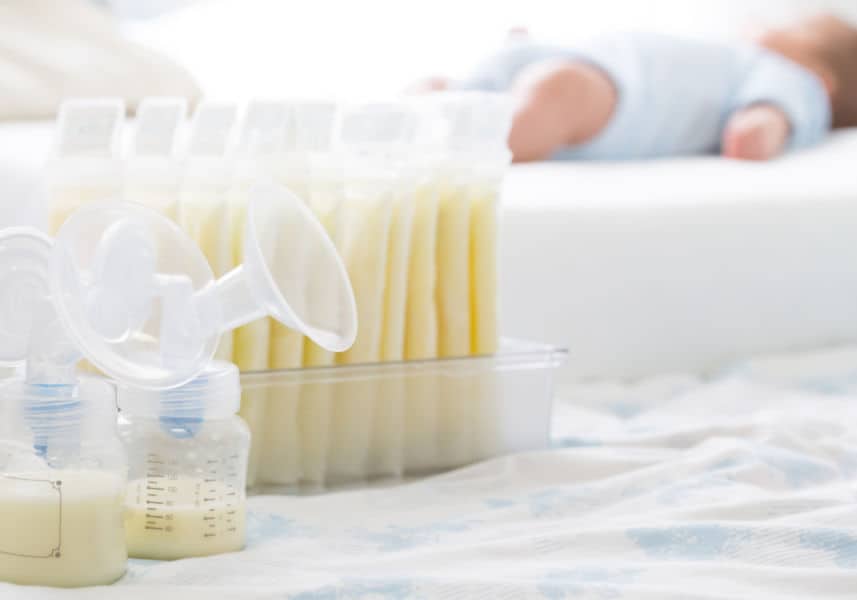 Image of breastmilk in freezer bags and breast pump parts nearby, with baby laying on bed in the background.