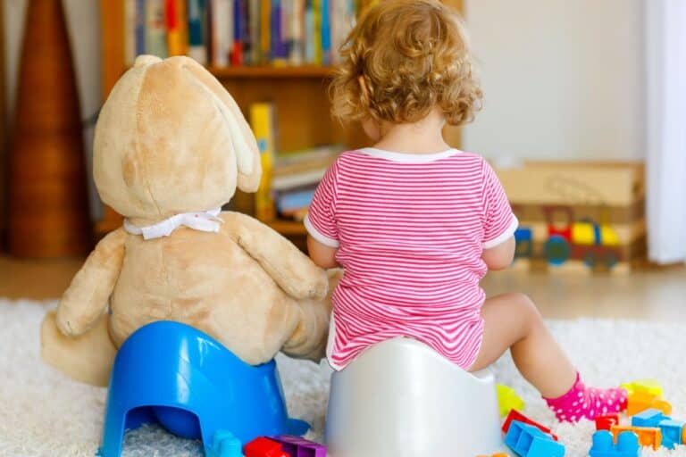 Stubborn toddler potty training in living room with toy next to her in potty chair too.