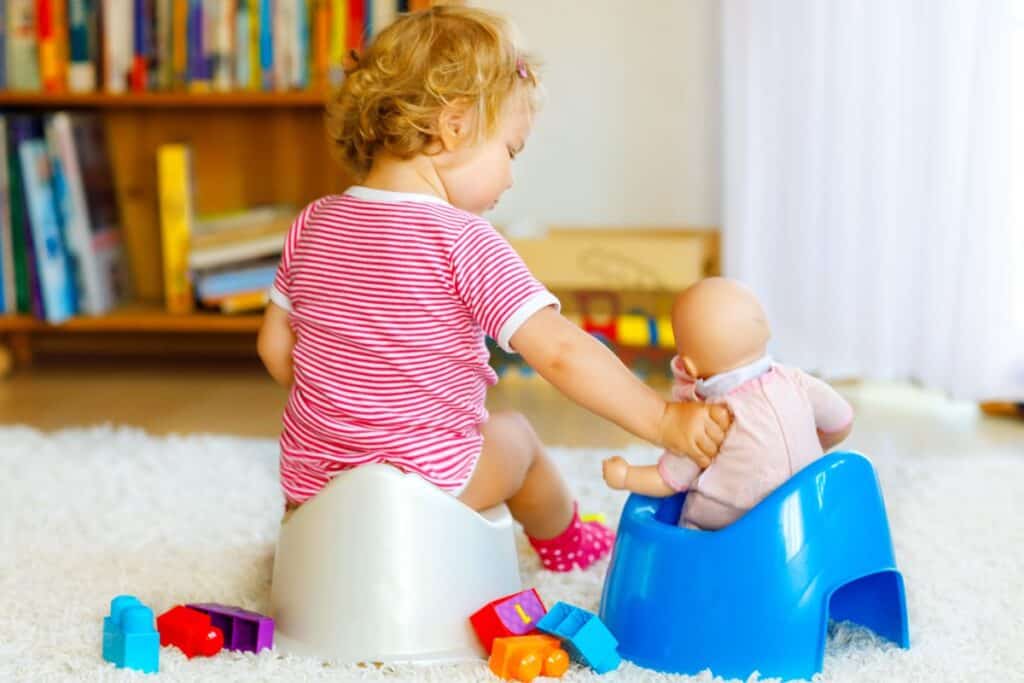 Toddler sitting on botty chair beside a doll sitting on potty chair.