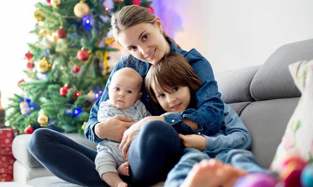 Christmas Eve traditions: mom cuddling young boy and a baby on a couch with Christmas tree in the background.