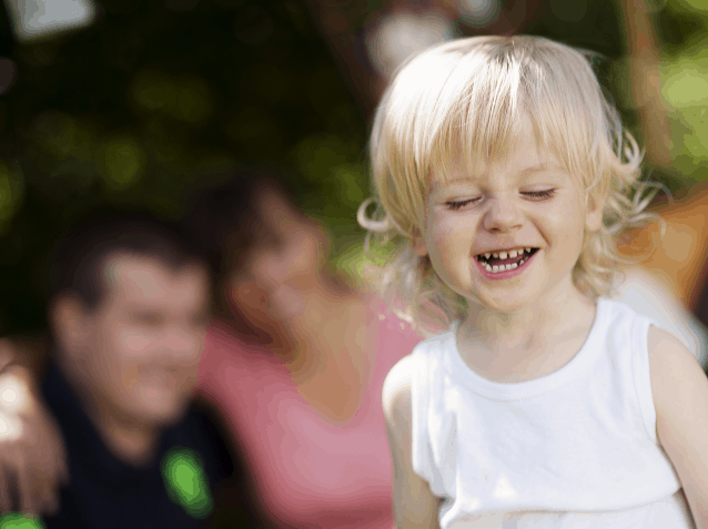 Mom and dad in background watching toddler play outside. Concept of Making time to make memories while watching their preschool daughter play outside.