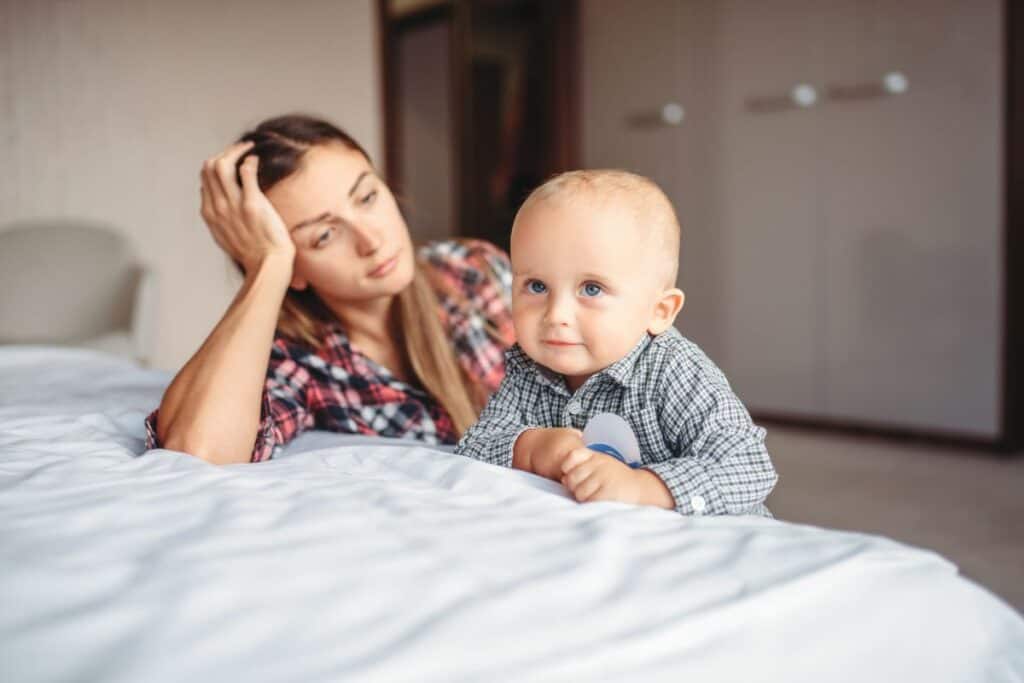 mom feeling sad, sitting against bed with baby boy.