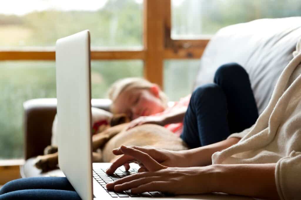 mom typing on laptop with sleeping child on couch beside her.