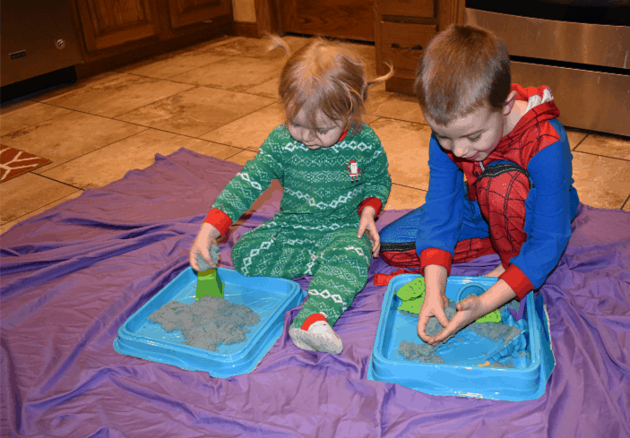 Image of a young boy and girl sitting on a purple sheet on the kitchen floor, playing with moldable Kinetic Sand: concept of Indoor Activities Ideas for Kids