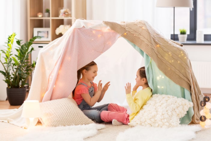 Two girls playing inside a homemade Indoor fort with fairy lights- concept of activity idea for kids at home.