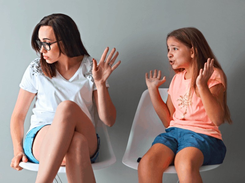 Mother and daughter sitting in chairs with animated, upset daughter and mom holding up her hand; talking back behavior