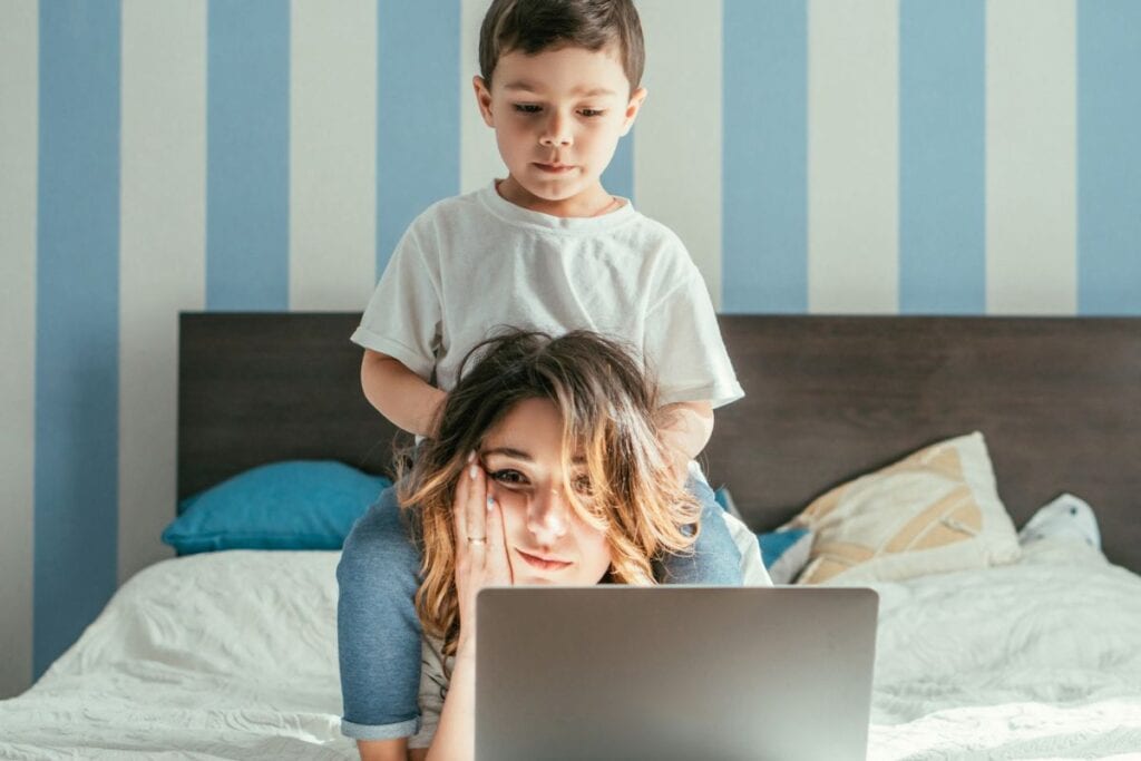 Mom sitting on bed with laptop, looking defeated with toddler sitting on her back.