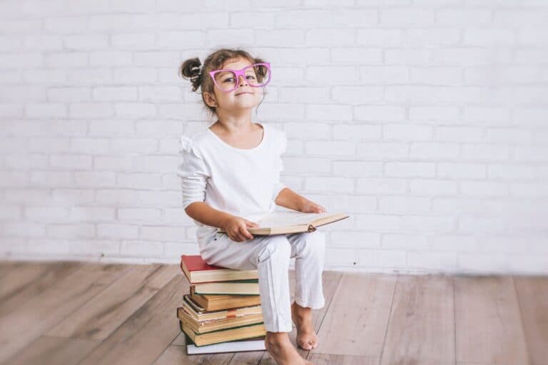 Young girl sitting on a stack of books with pretend glasses.