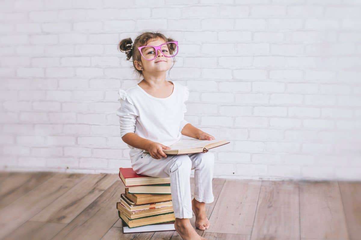 Young girl sitting on a stack of books with pretend glasses.