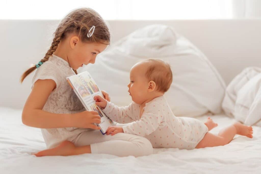 Little girl with braids sitting on a bed with baby sister, reading her a book.