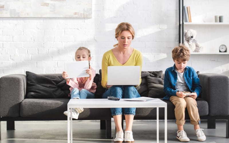 image of mother and children using different electronic devices side by side on a couch in the living room, ignoring each other.