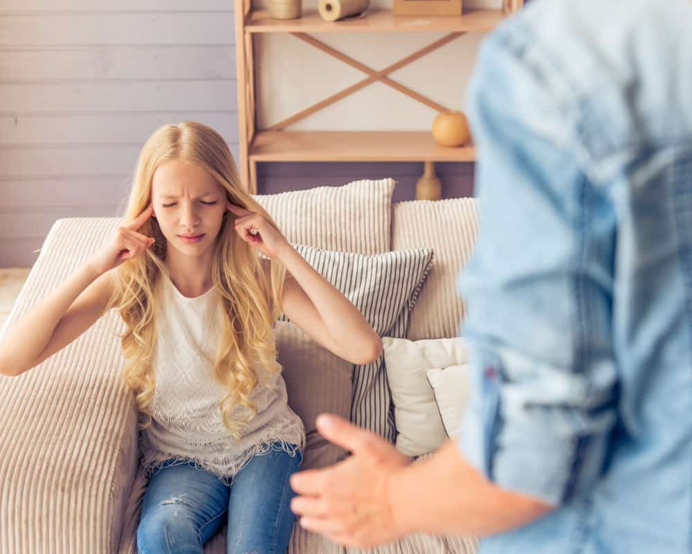 Image of tween girl sitting on couch with fingers in her ears and eyes closed with mother standing in front of her, exasperated and trying to reason with her daughter. Concept of tween discipline- de-escalating tween drama.