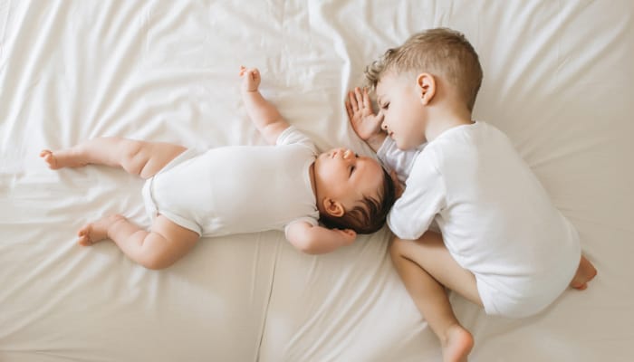 Toddler boy crouched laying on a bed beside a newborn baby: example of potty training setbacks with new baby