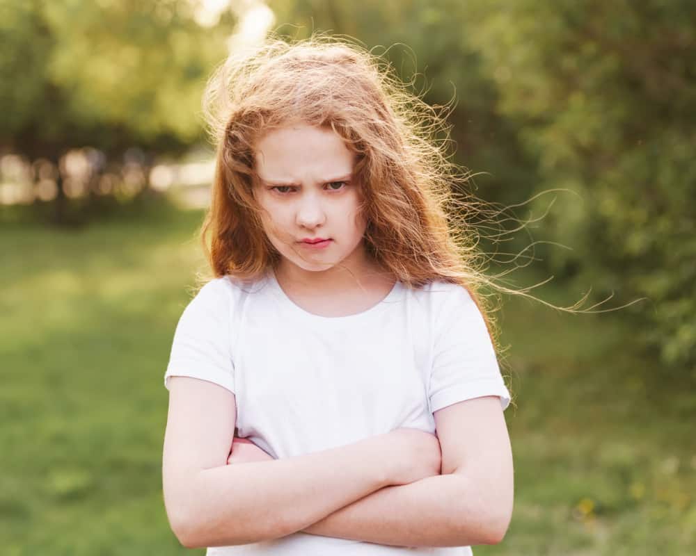 Image of a young girl standing outside, frowning with her arms crossed; preteen emotions- handling girl drama