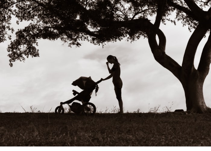 Woman standing under a tree with a baby stroller, stopping to gather her thoughts.