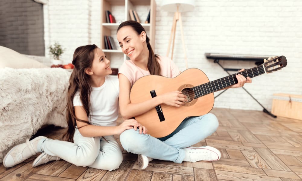 playing guitar - bored stay at home mom ideas: a mom playing an acoustic guitar on the floor with her preteen daughter next to her smiling.