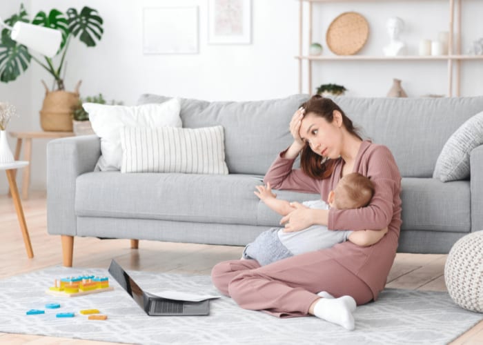 Mom leaning against couch, holding baby in one arm and looking into distance- concept of moms do so much and face burnout as a result.