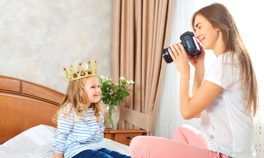 housewife hobbies- photography: a young mom uses a digital camera to take a snapshot of her toddler sitting on a bed with a princess crown on her head.