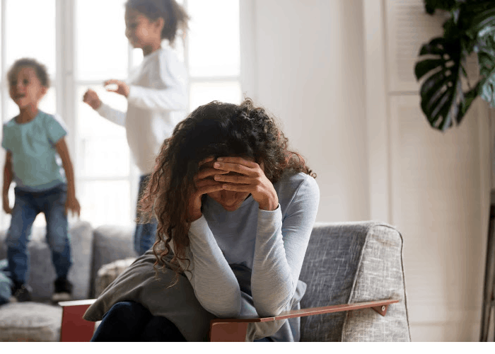 A mom with her head in her hands with kids fighting in the background.