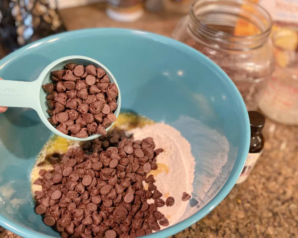 Pouring a cup full of chocolate chips into a mixing bowl with cake mix and butter.