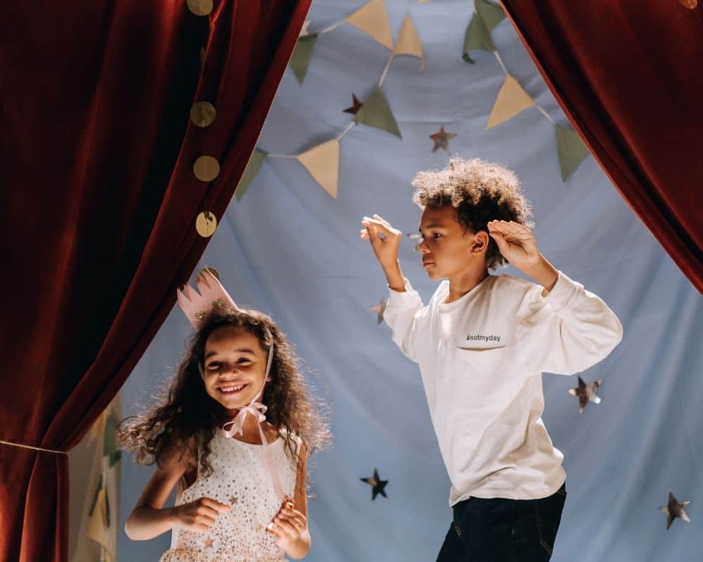 image of a brother and sister acting out a play in front of a makeshift curtain with gold banners hanging behind them. Concept of indoor activities for all ages to participate in.