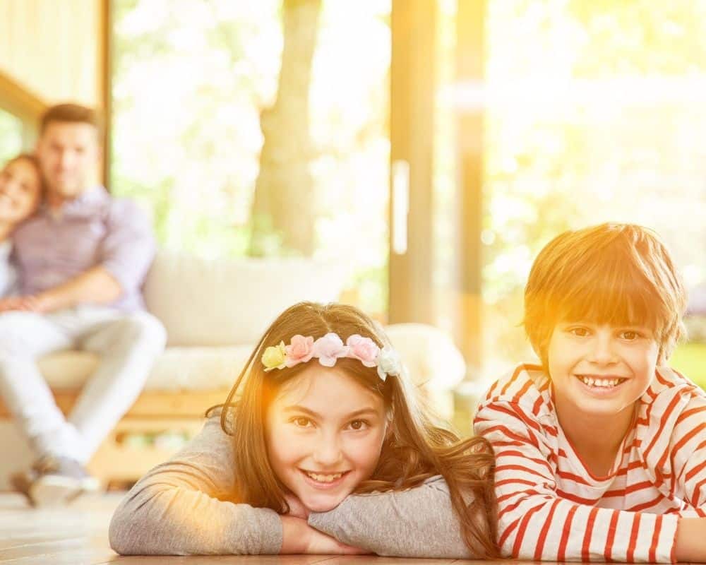 Image of brother and sister preteens side by side on living room floor, smiling with parents on couch in background. 