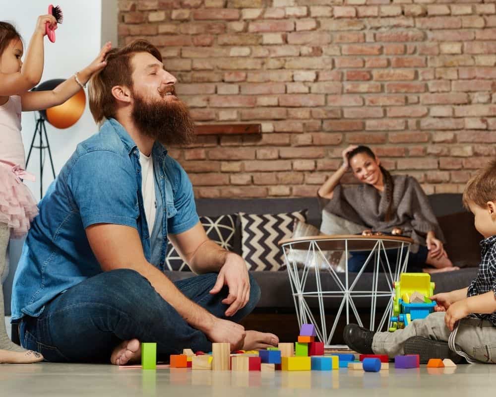 Image of Dad playing on living room floor while toddler son plays with building blocks and preschool aged daughter combs his hair with a play comb. Concept of screen-free family fun.
