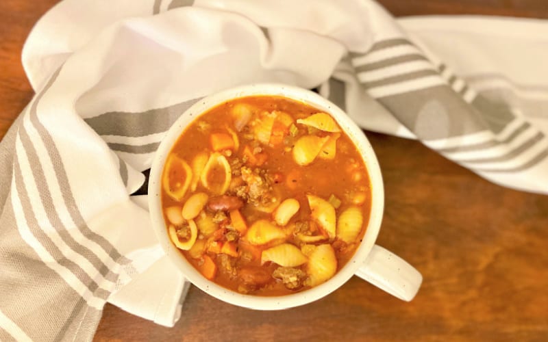 Image of bowl of pasta e fagioli soup on a brown table with a white and grey striped hand towel surrounding it.