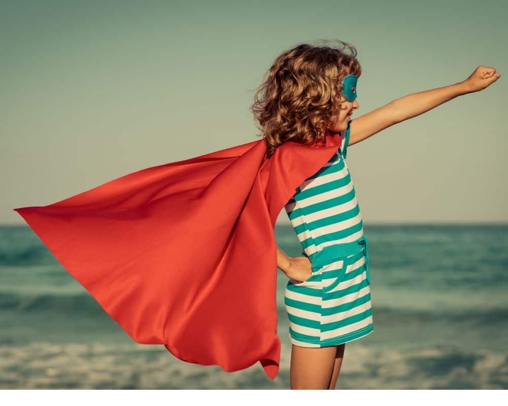 Image of girl standing on beach, wearing a red cape flowing in the wind. Concept of raising a leader.