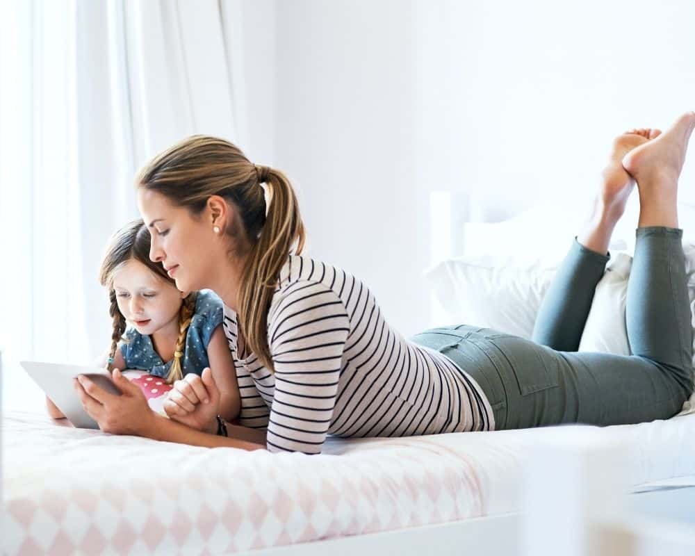 Image of young mom and grade school aged daughter laying on child's bed, watching a TV show on child's tablet.