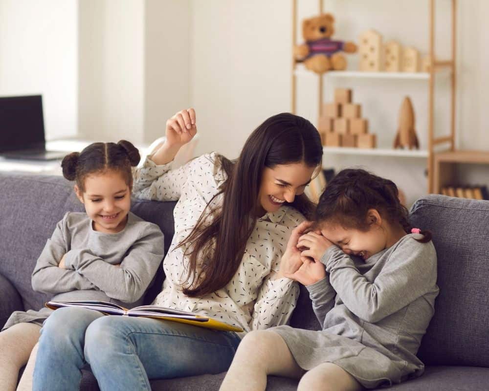 Image of mom and twin daughters sitting on a couch at home, reading a book and tickling - having fun connecting as a family.