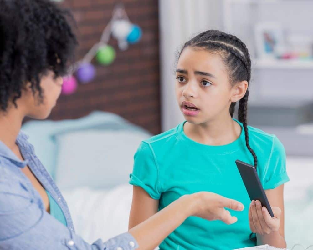 Mom holding out her hand motioning for teen daughter to hand her the smart phone, with daughter looking upset and argumentative.