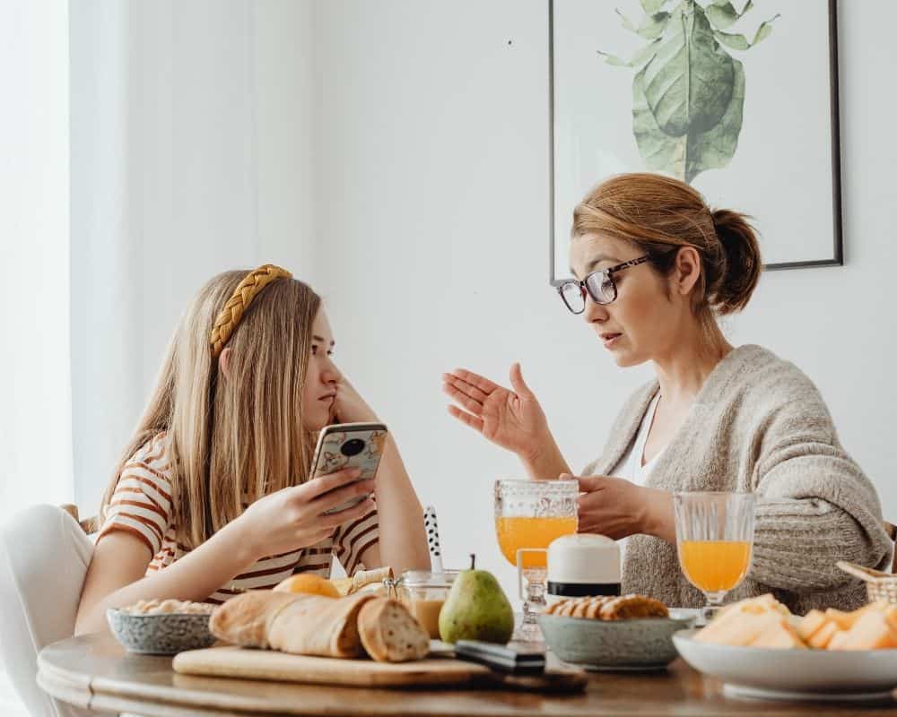 Image of mother and preteen daughter at breakfast table, with mother holding up her hand while explaining something to her daughter.