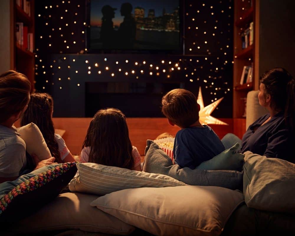 Image of mom, dad and three kids resting on pillows on the sofa, watching a family movie with twinkling lights in the background for ambiance.