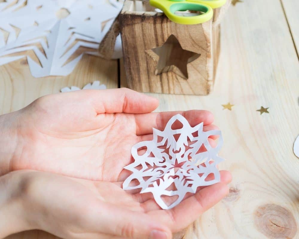 Image of a teen's hands holding a finished DIY paper snowflake over a craft table.
