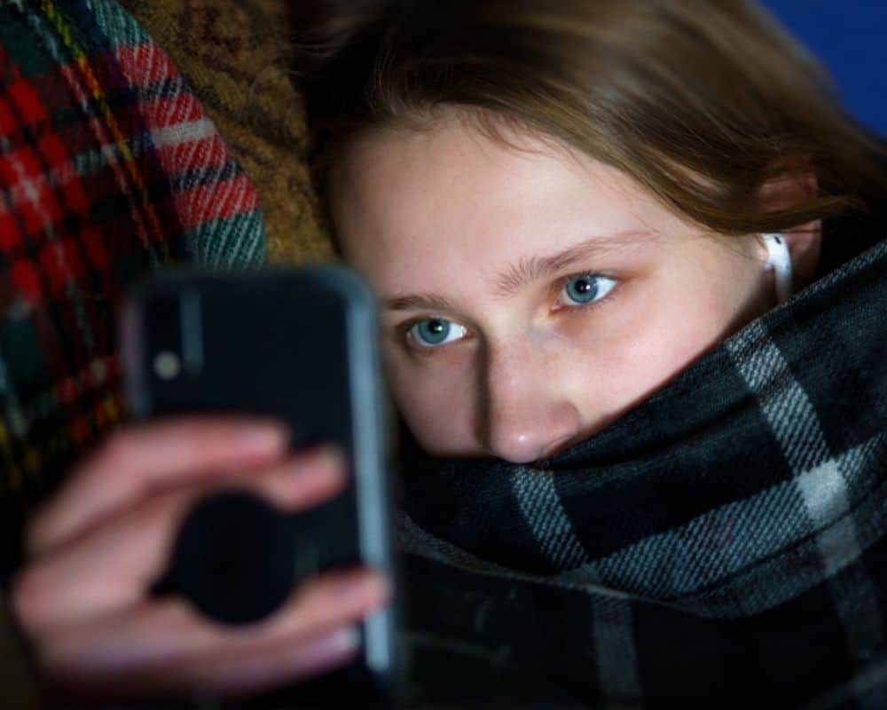 Image of teen girl huddled under blanket on couch with earbuds, staring aimlessly at cell phone. Concept of screen addiction.