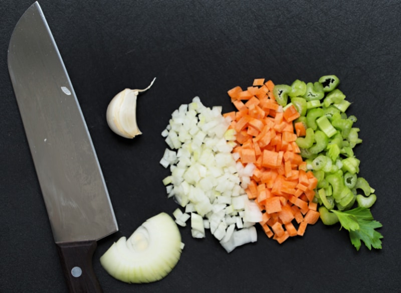 Image of chef's knife with diced onions, garlic, carrots and celery on a dark background.