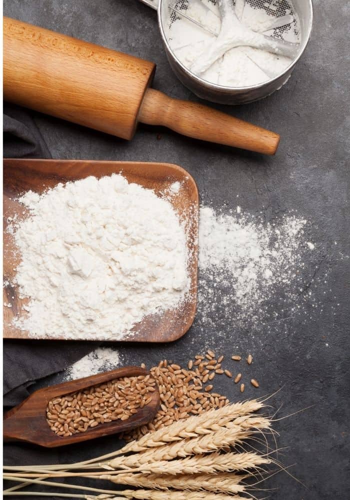 homemade white bread ingredients- flour, wheat berries and rolling pin on a black countertop.