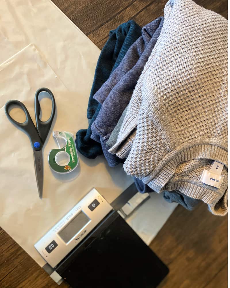 Image of a stack of clothing, a postage scale, tape and scissors on top of poly bags on a wooden table. Concept of preparing items to ship on Marketplace.