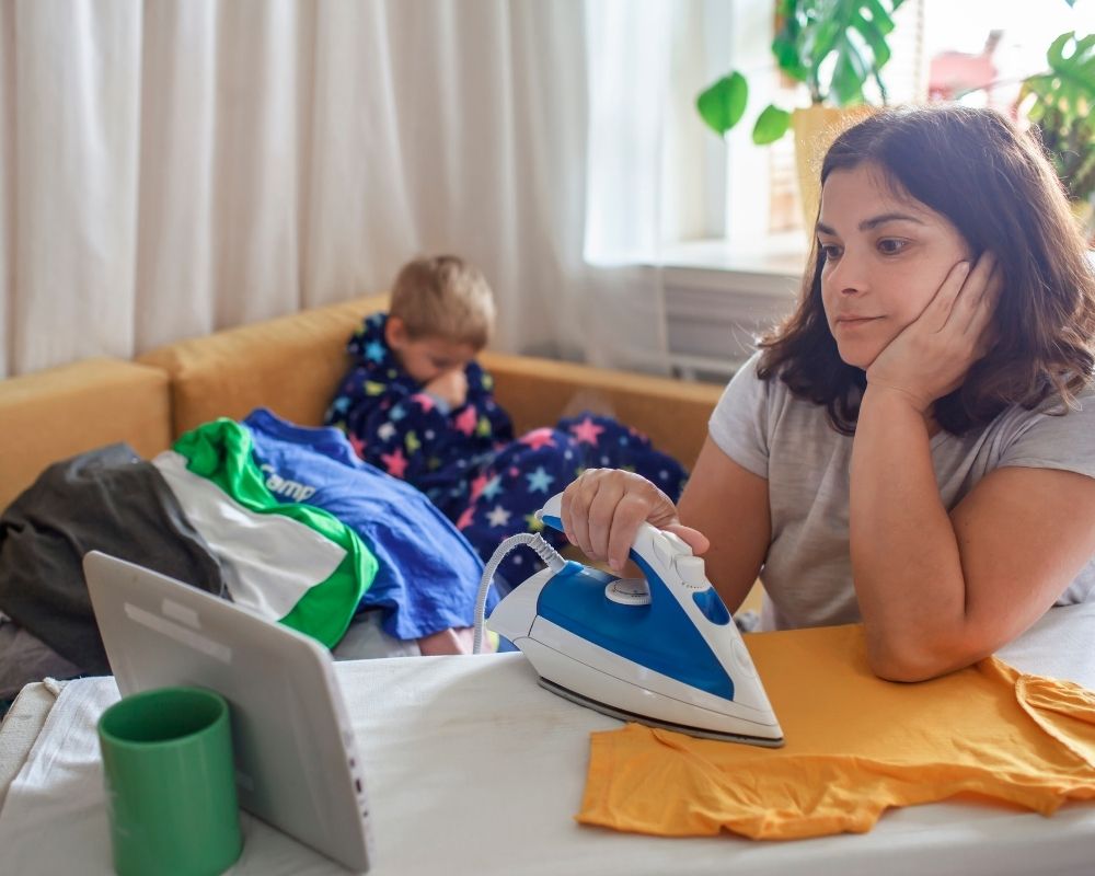 Mom ironing, distracted and watching a tablet with kid sitting on couch in background.