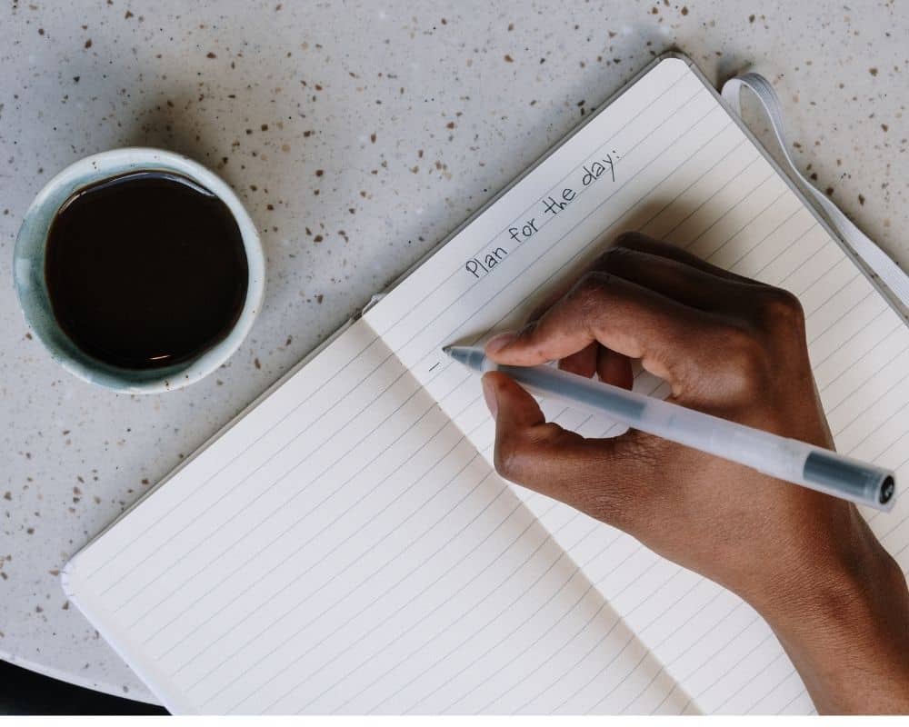 Image looking down at a woman's hand writing on a notebook -Plan for the day- with a cup of coffee beside it. Concept of making plans and goals in the New Year.