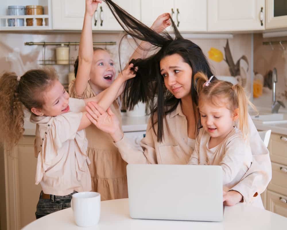 Image of a mom with three daughters surrounding her at the kitchen table, playing with her hair, being wild and mom looking calmly bewildered. Concept of the craziness of having four kids.