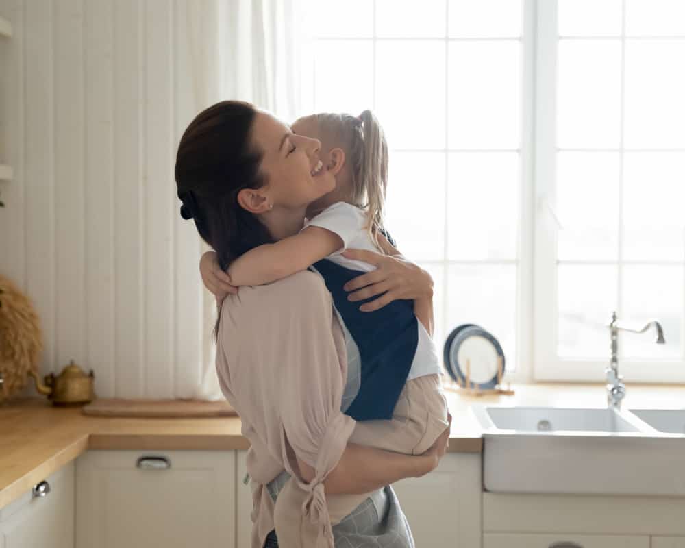 Mom in a bright white kitchen smiling and holding her toddler in her arms; concept of relaxed mom- the chores can wait.
