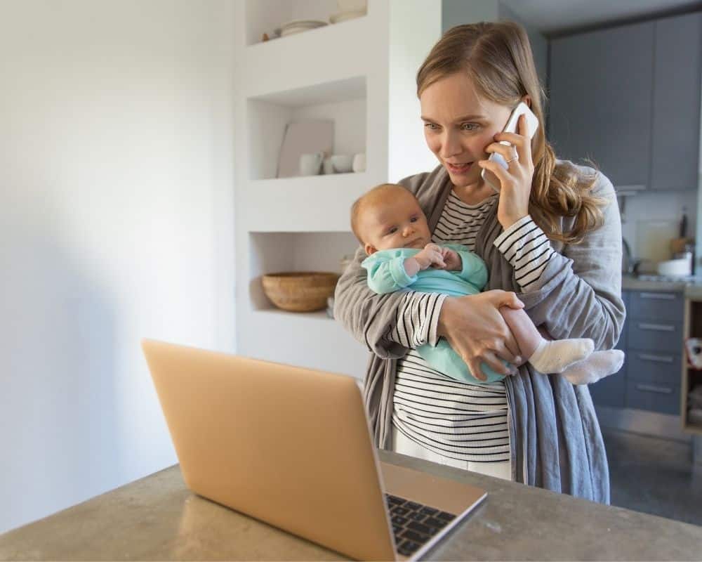 Image of worried mom holding a newborn baby, looking at a laptop screen and talking on a phone. 