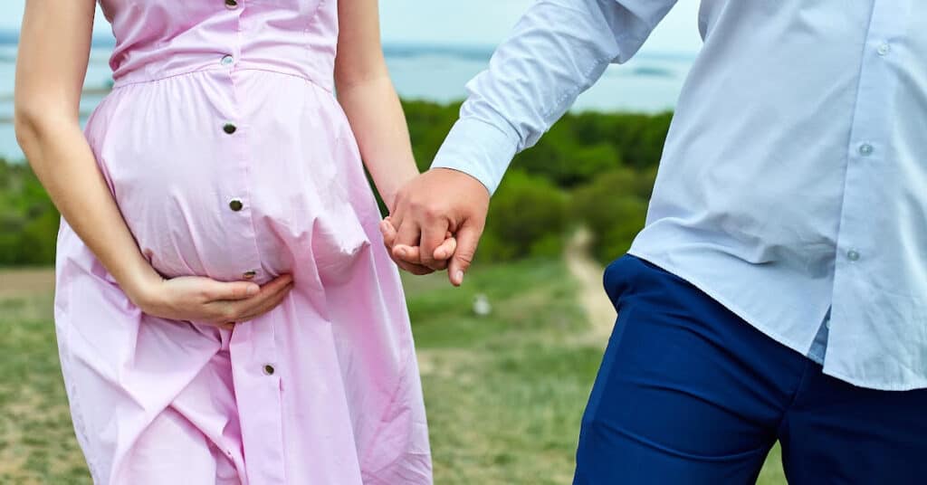 Image of lower half of woman standing outside resting her hand underneath her pregnant belly, holding the hand of her husband. Concept of planning a large family.