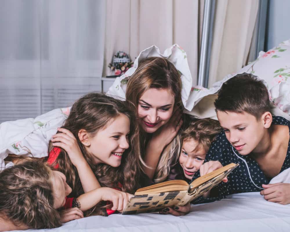 Image of a mom and her four kids laying on a bed and reading a book together.