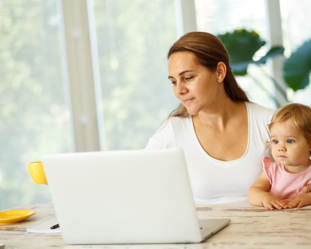 Woman working on laptop while holding baby and drinking coffee.